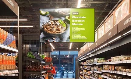 Aan het plafond van een supermarkt hangt een groot scherm.