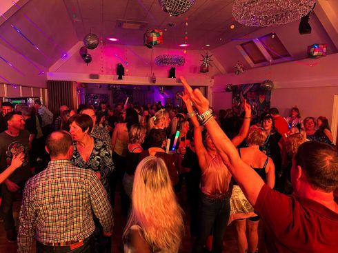 A crowded dance floor at a party with disco balls, pink lighting, and people dancing with their arms raised.