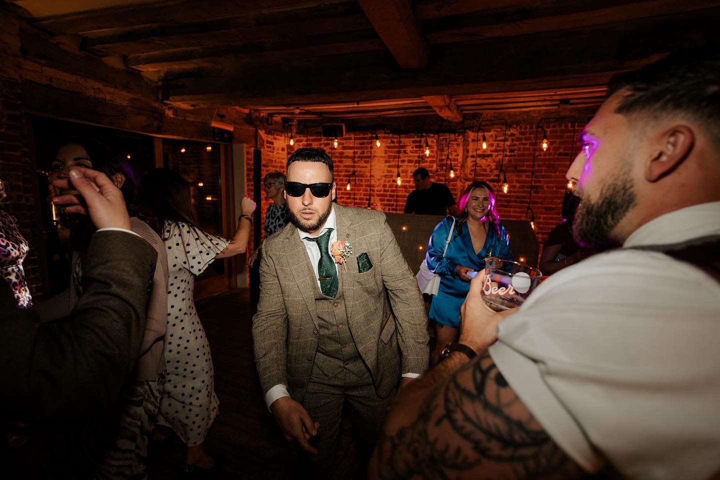A person wearing sunglasses and a patterned suit dances at a party in a dimly lit, rustic room with brick walls.