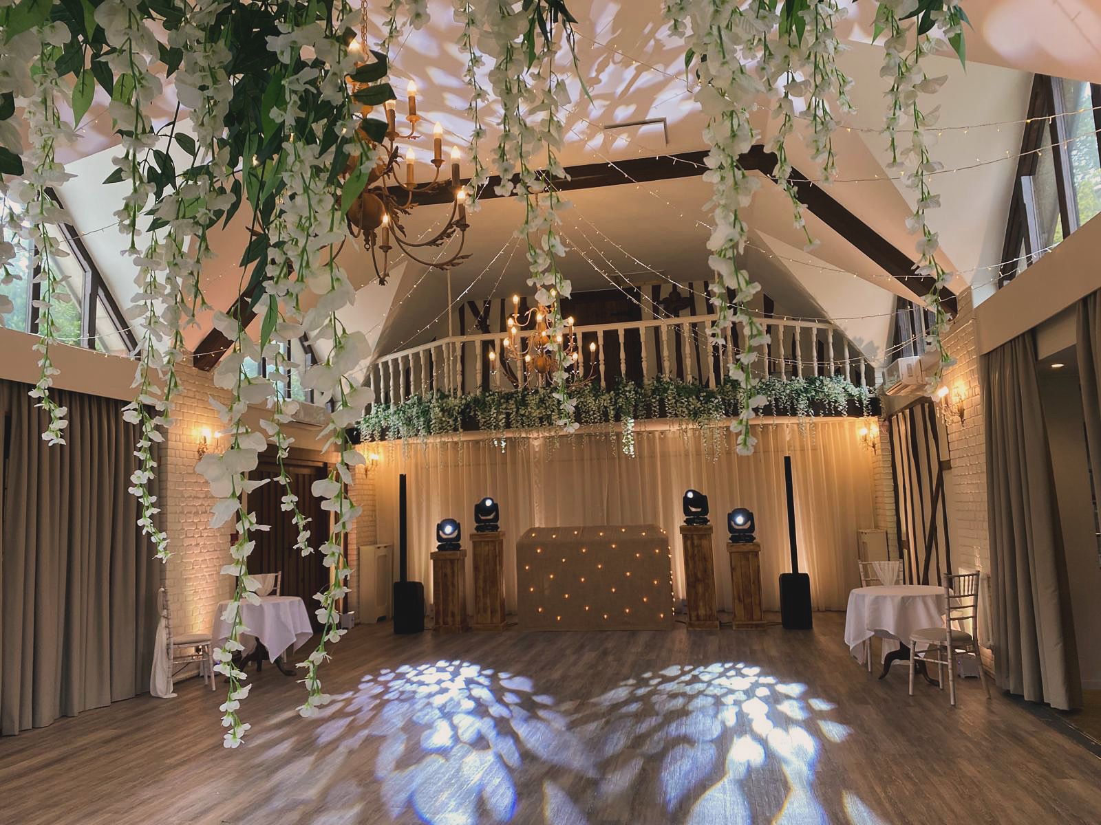 An indoor wedding reception venue with hanging white floral vines, a stage, and decorative floor lighting.
