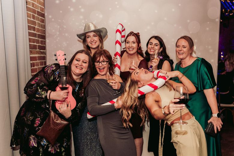 Seven friends posing at a holiday party, some holding a candy cane prop and wine glasses, smiling in a festive setting.