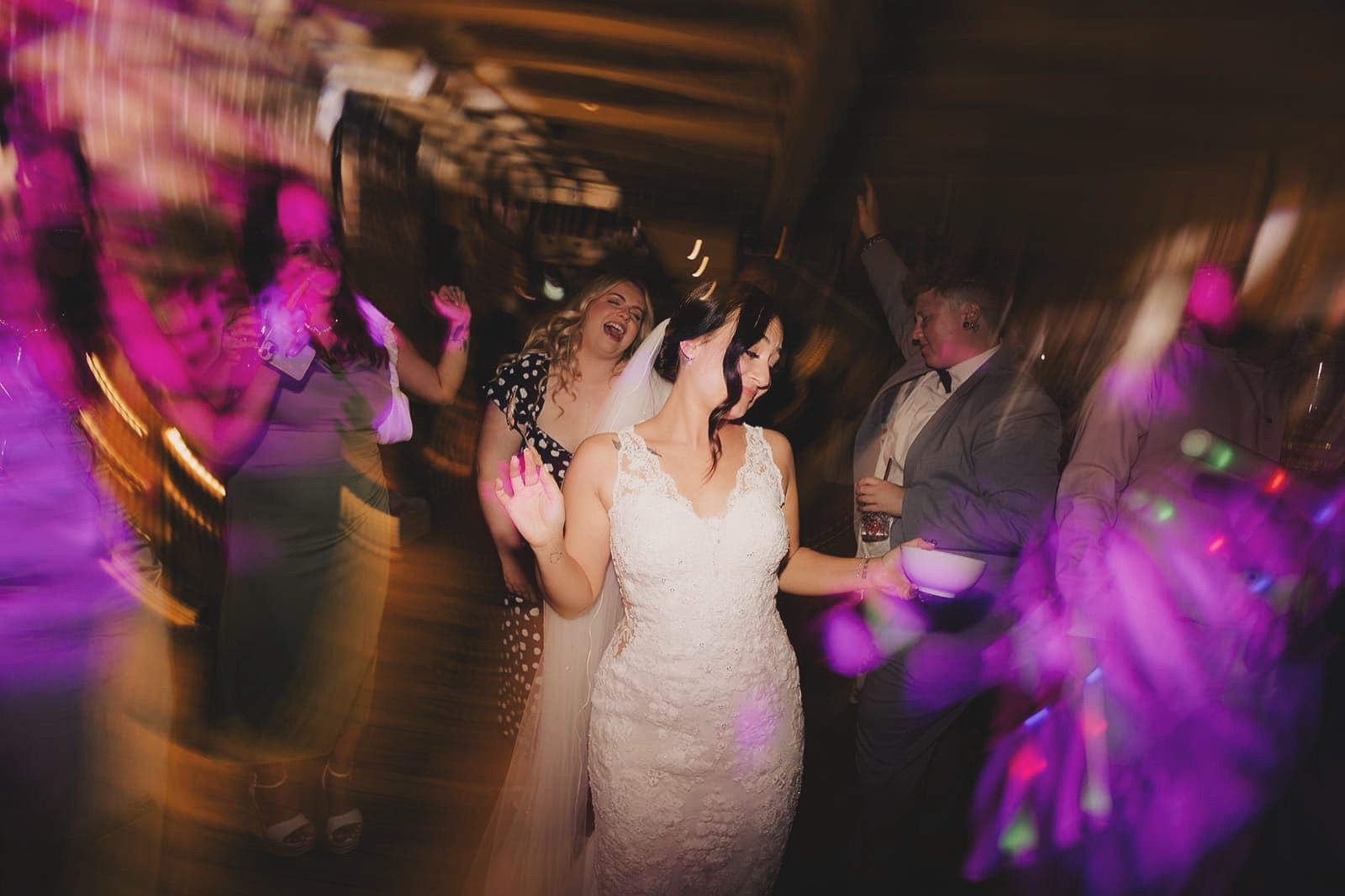 A bride dancing in a white lace dress, surrounded by guests at a dimly lit, motion-blurred party with colorful lights.