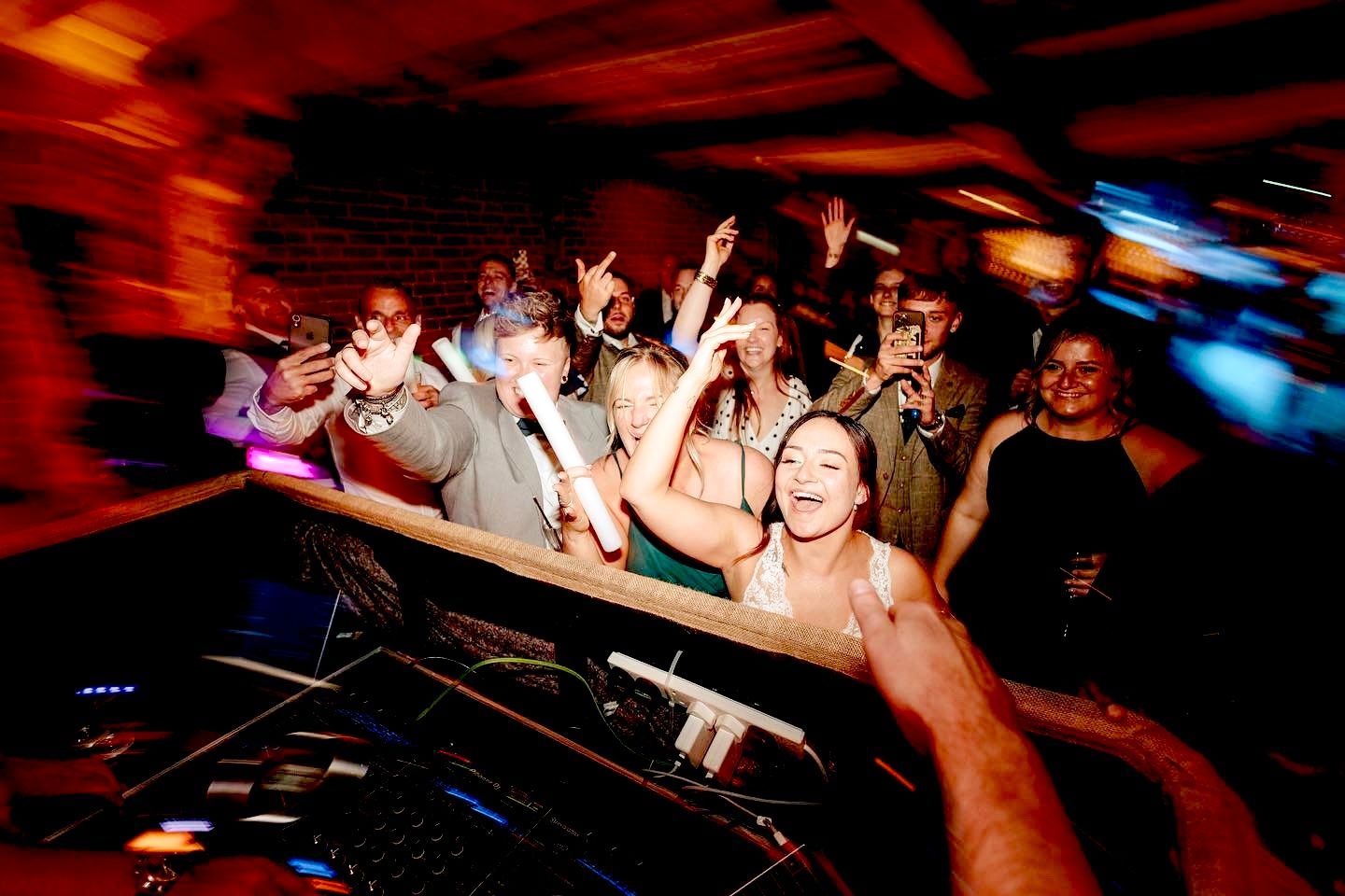 A crowded, energetic dance floor at an event, with people dancing and raising their arms under warm, dynamic lighting.