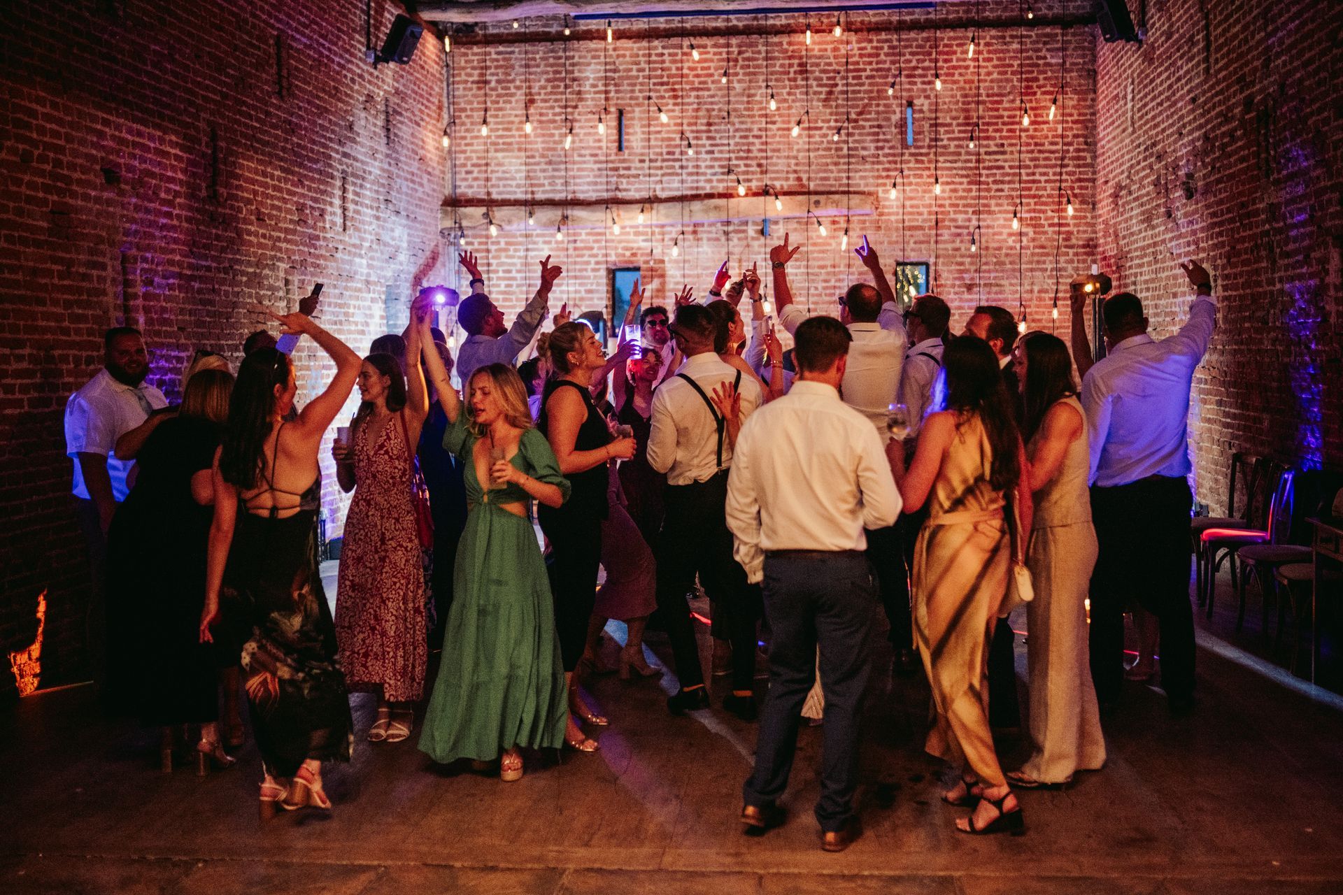 A festive group of people dancing together in a rustic brick event hall illuminated by string lights.