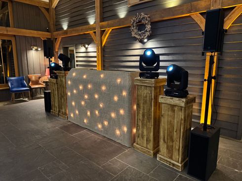 A DJ booth with a lit-up front panel, wooden pillars, and stage lighting equipment, set up in a wooden-walled room.