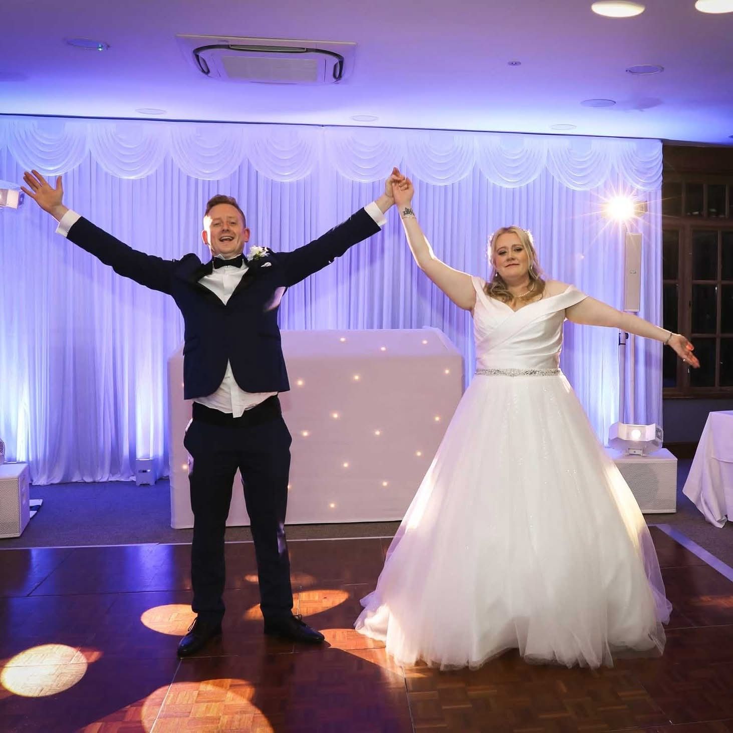 A bride and groom in wedding attire stand with arms outstretched in celebration on a dance floor with stage lighting.