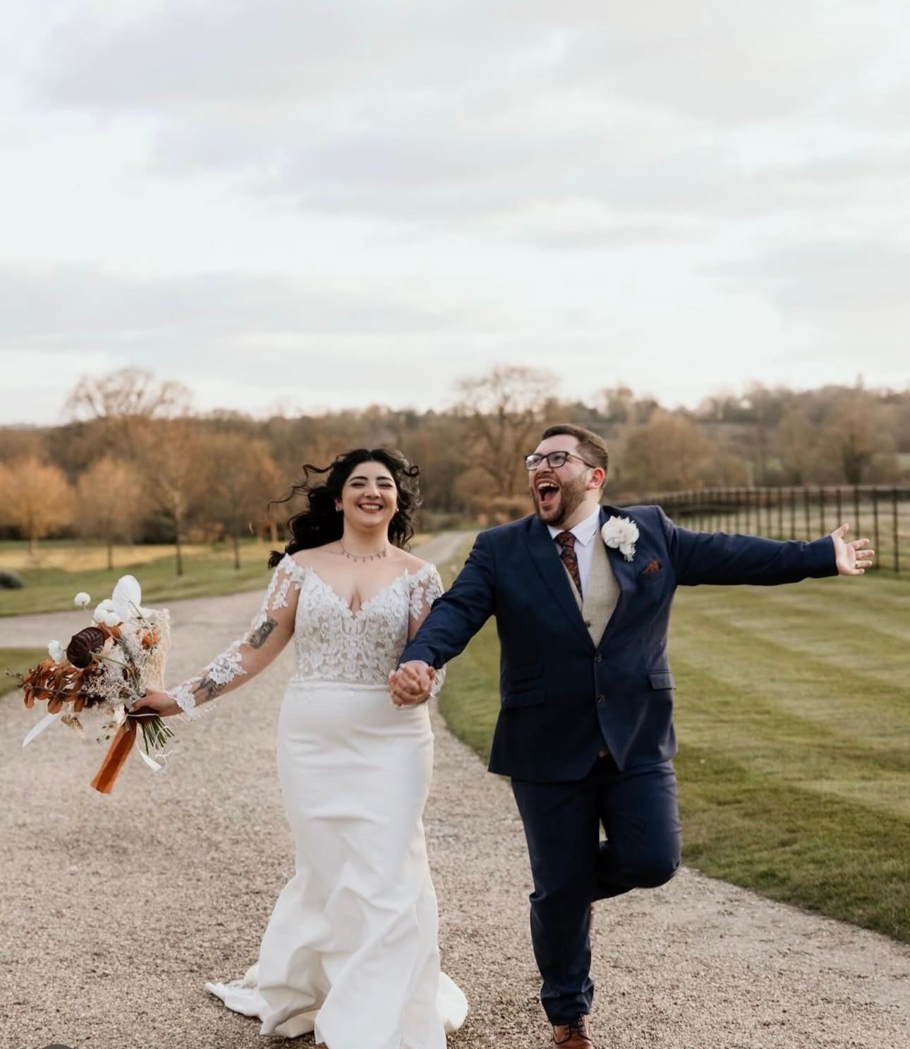 A joyful couple running hand-in-hand down a gravel path at an outdoor wedding, holding a bouquet with arms outstretched.