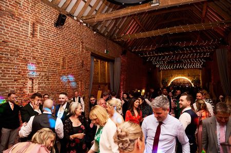 A wedding reception inside a rustic brick barn with string lights, guests socializing and dancing.