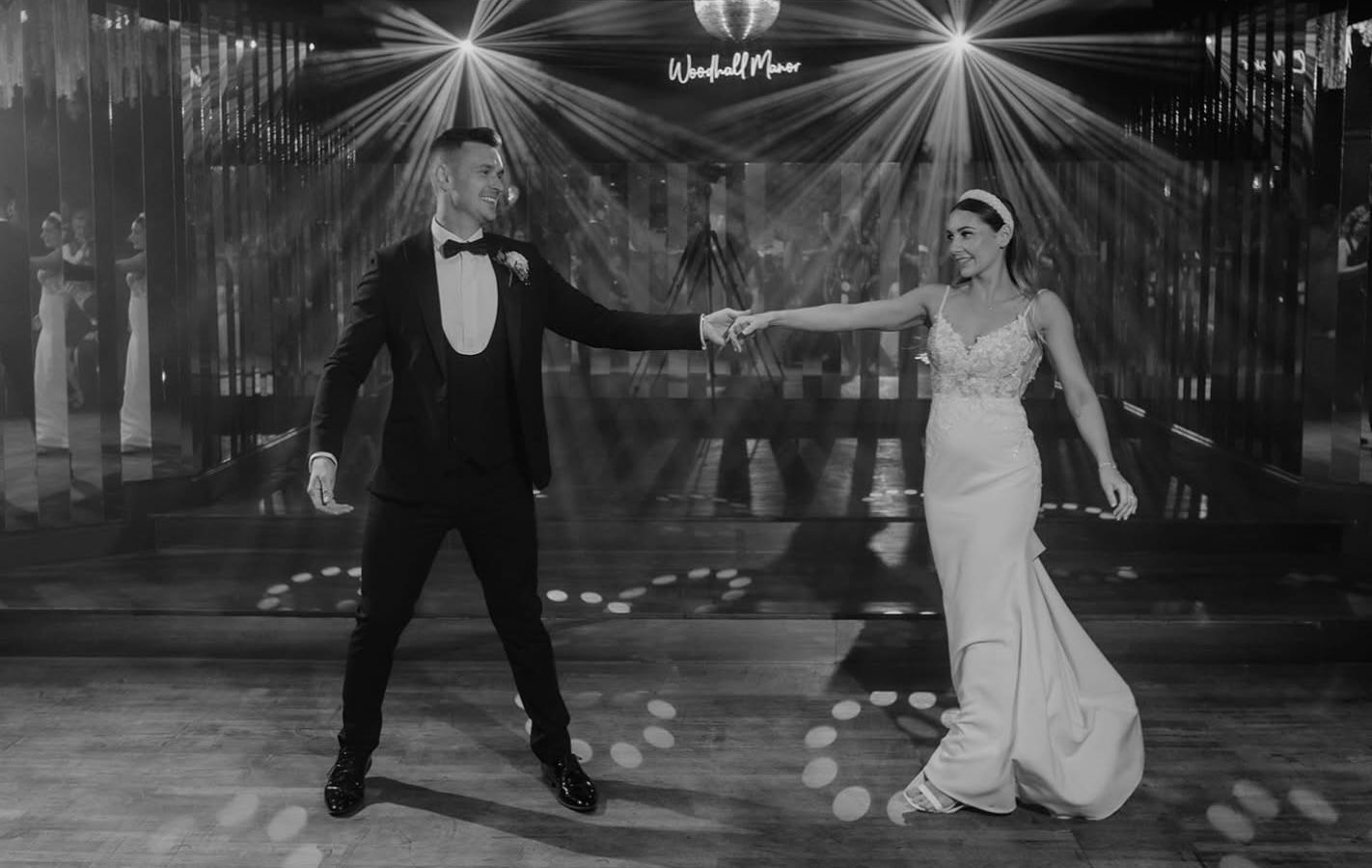 A bride and groom in formal attire dance together under stage lights at a wedding reception.