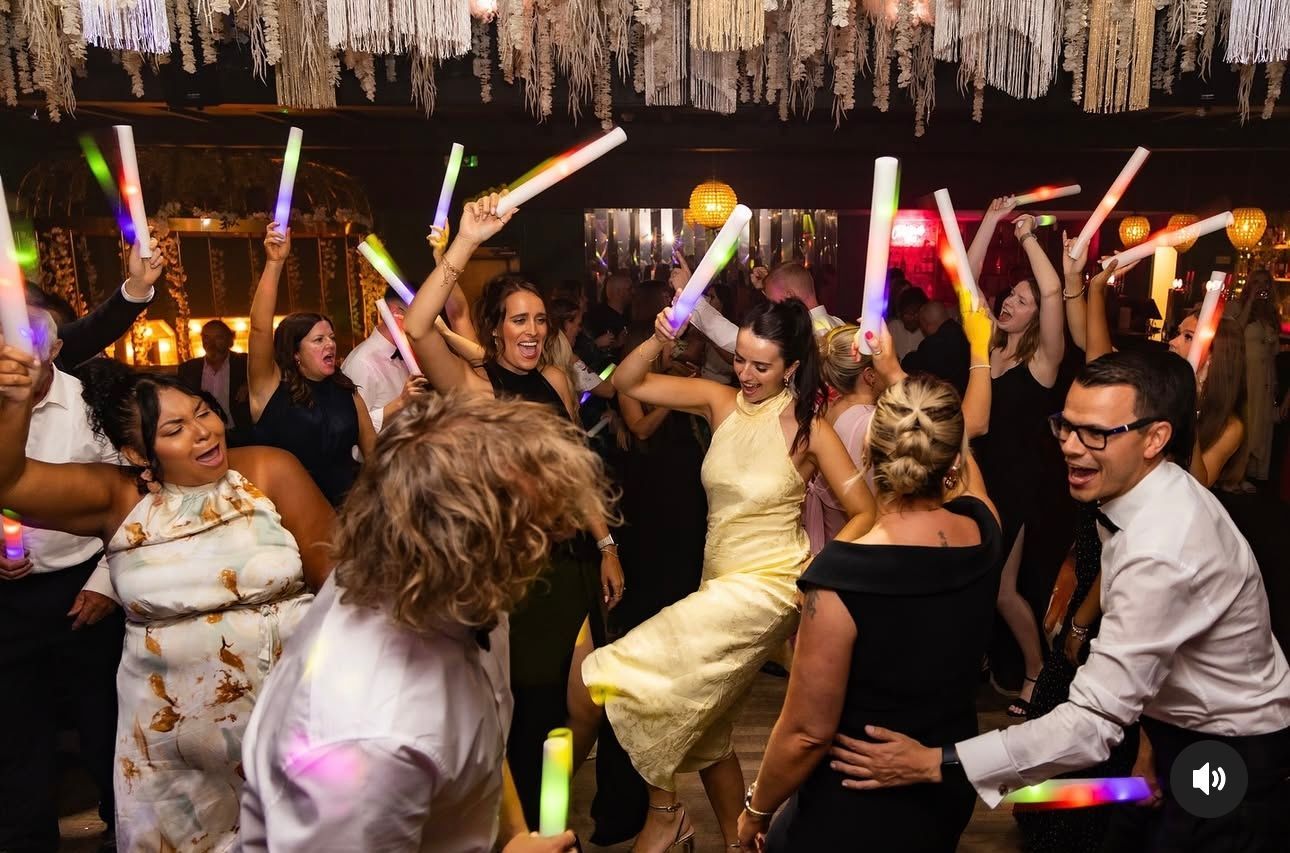 A crowd dances in a decorated venue, waving glowing light sticks in the air during a celebration.