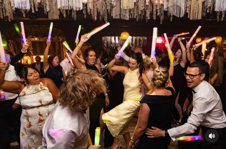 A crowd dances in a decorated venue, waving glowing light sticks in the air during a celebration.