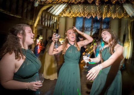 Three people in long green dresses dance and hold glasses in a decorated indoor venue with string lights.