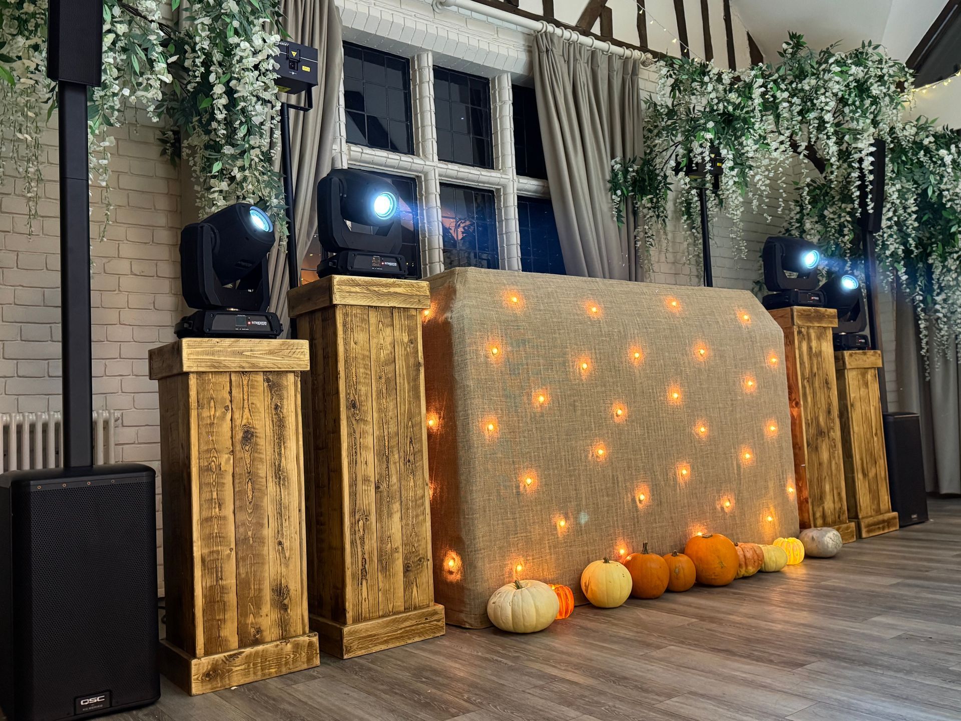 A rustic wedding or party DJ setup with illuminated burlap, wooden pedestals, stage lights, and pumpkins on the floor.