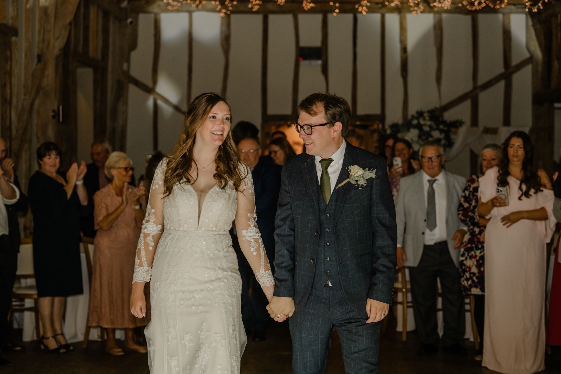 A wedding couple walks hand-in-hand through a rustic barn reception, smiling at guests while wearing formal attire.