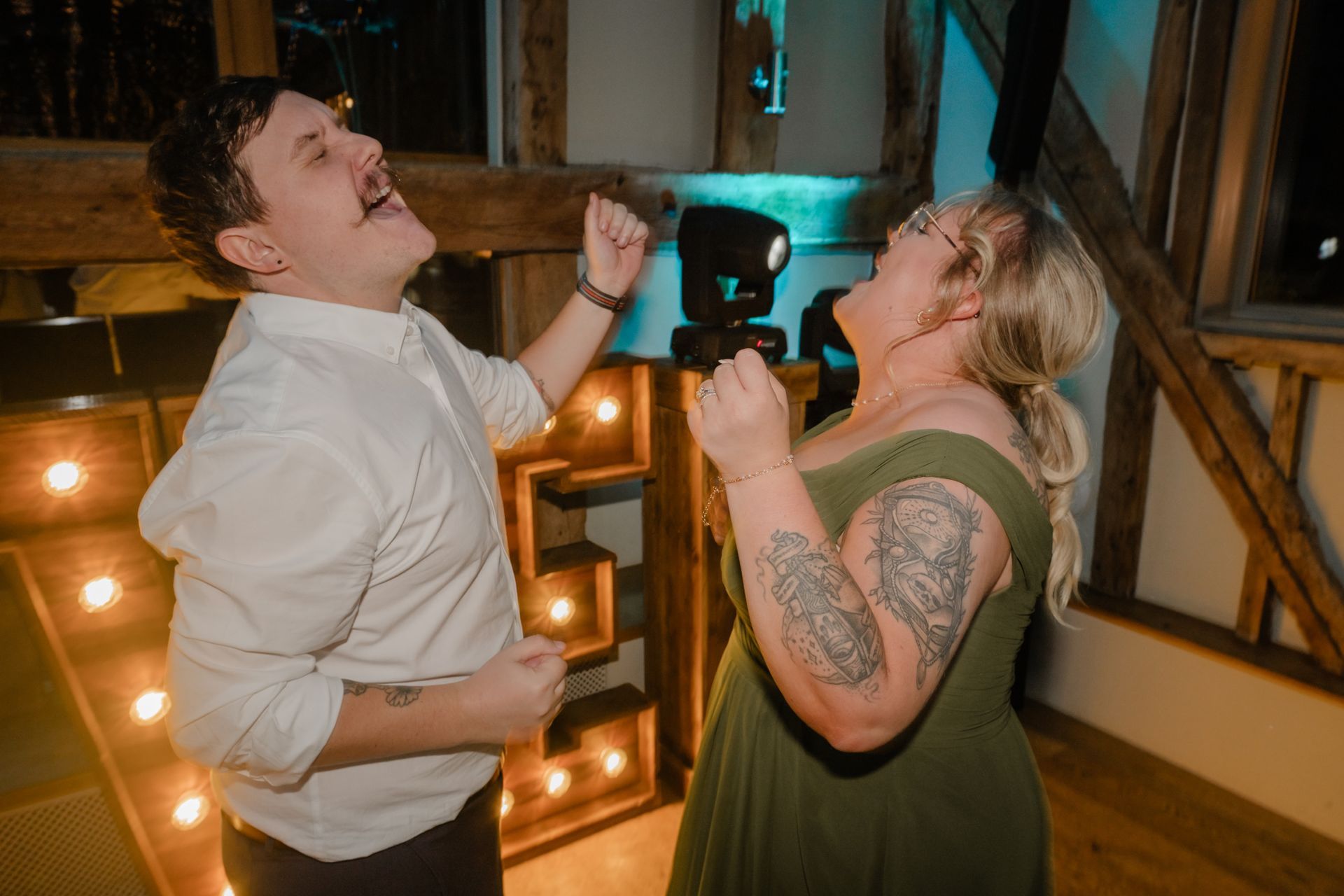 Two people dancing and singing with joy in a rustic, dimly lit indoor venue illuminated by marquee lights.