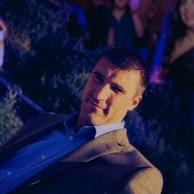 A man in a blue button-down shirt and a textured blazer looks toward the camera in a dimly lit, possibly indoor setting.