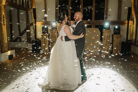 A bride and groom in wedding attire dancing in a rustic hall as white confetti falls around them.