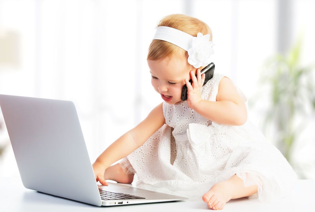 A toddler sits at a desk using a laptop and cell phone, wearing a white dress and headband.