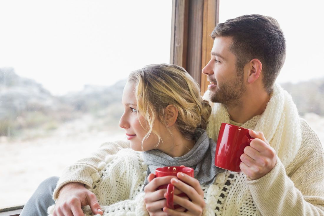 Couple gazing out a window, holding red mugs, wrapped in sweaters. Snowy outdoor background.