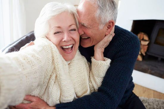 Elderly couple laughing, embracing. Woman in cream sweater, man in blue sweater near fireplace.