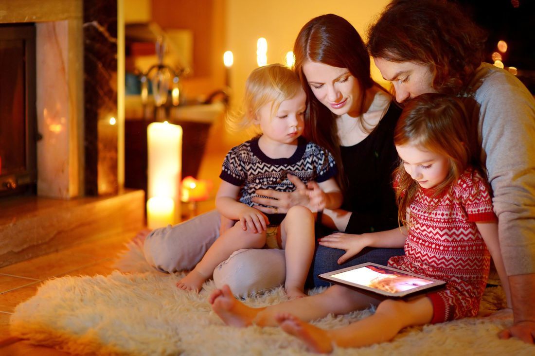 Family gathered by fireplace, looking at tablet. Candles provide warm glow.