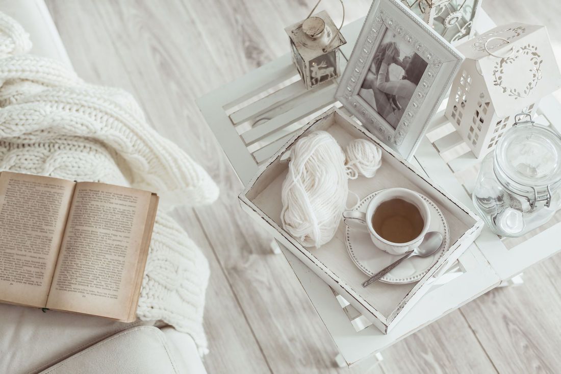 Cozy scene: Book, tea cup on tray with yarn, photo frame, and lantern on a white wooden table.
