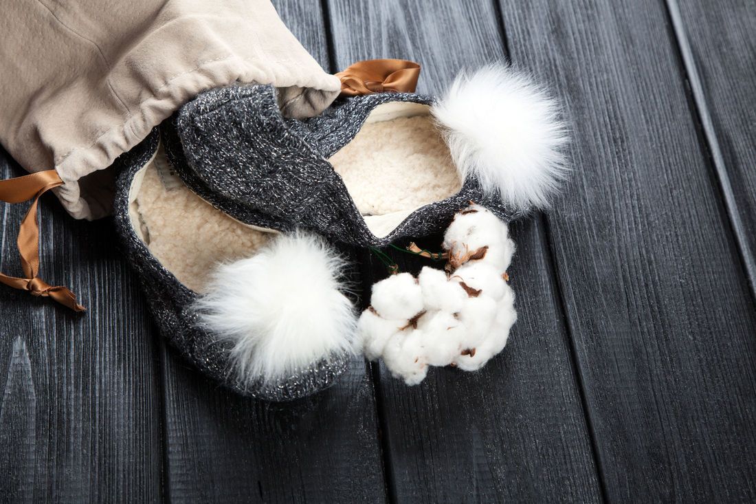 Gray slippers with white pom-poms and cotton sprig spilling from a tan drawstring bag on a dark wood surface.