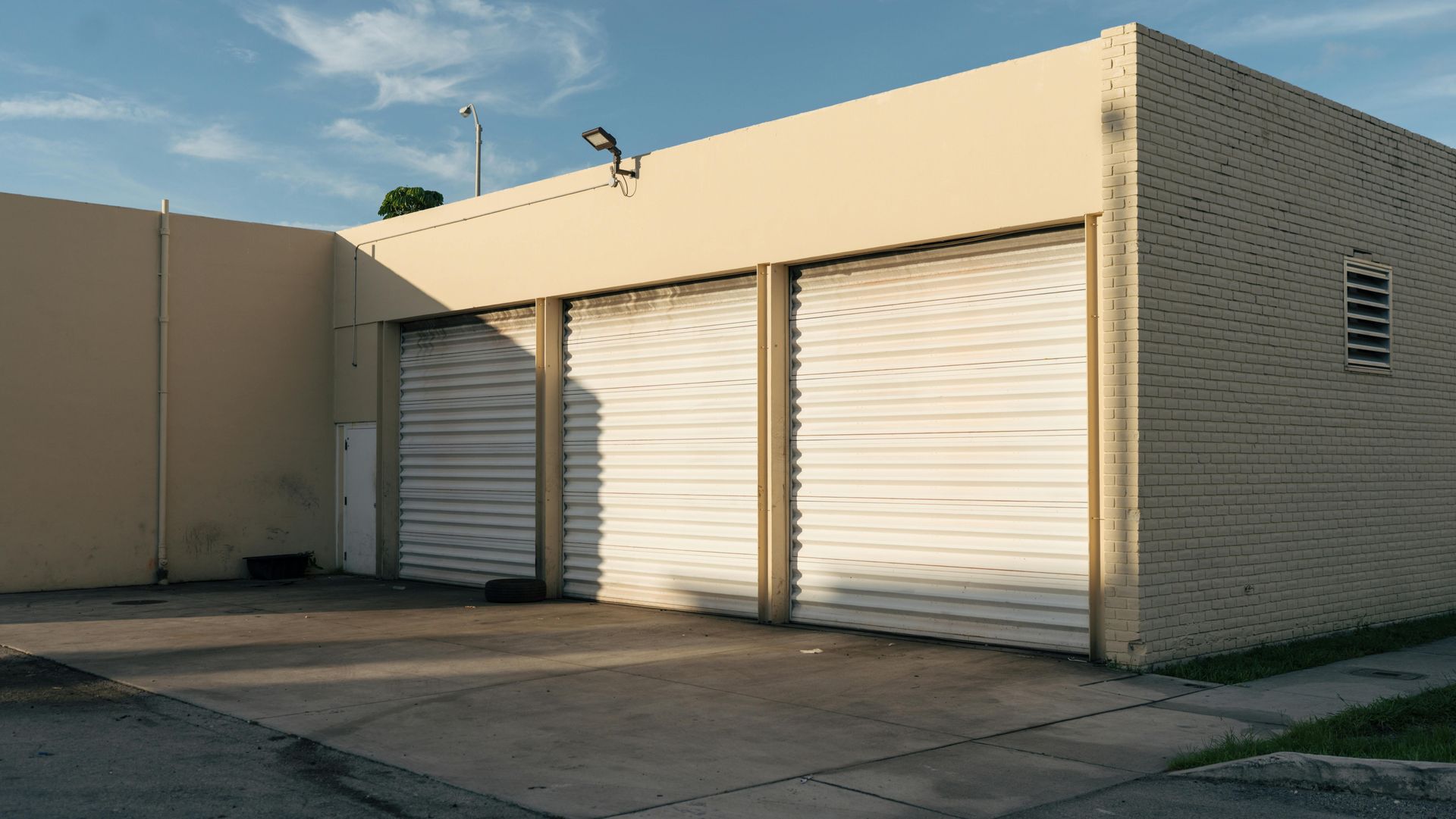 Exterior of a tan building with closed garage doors on a concrete driveway under a blue sky.