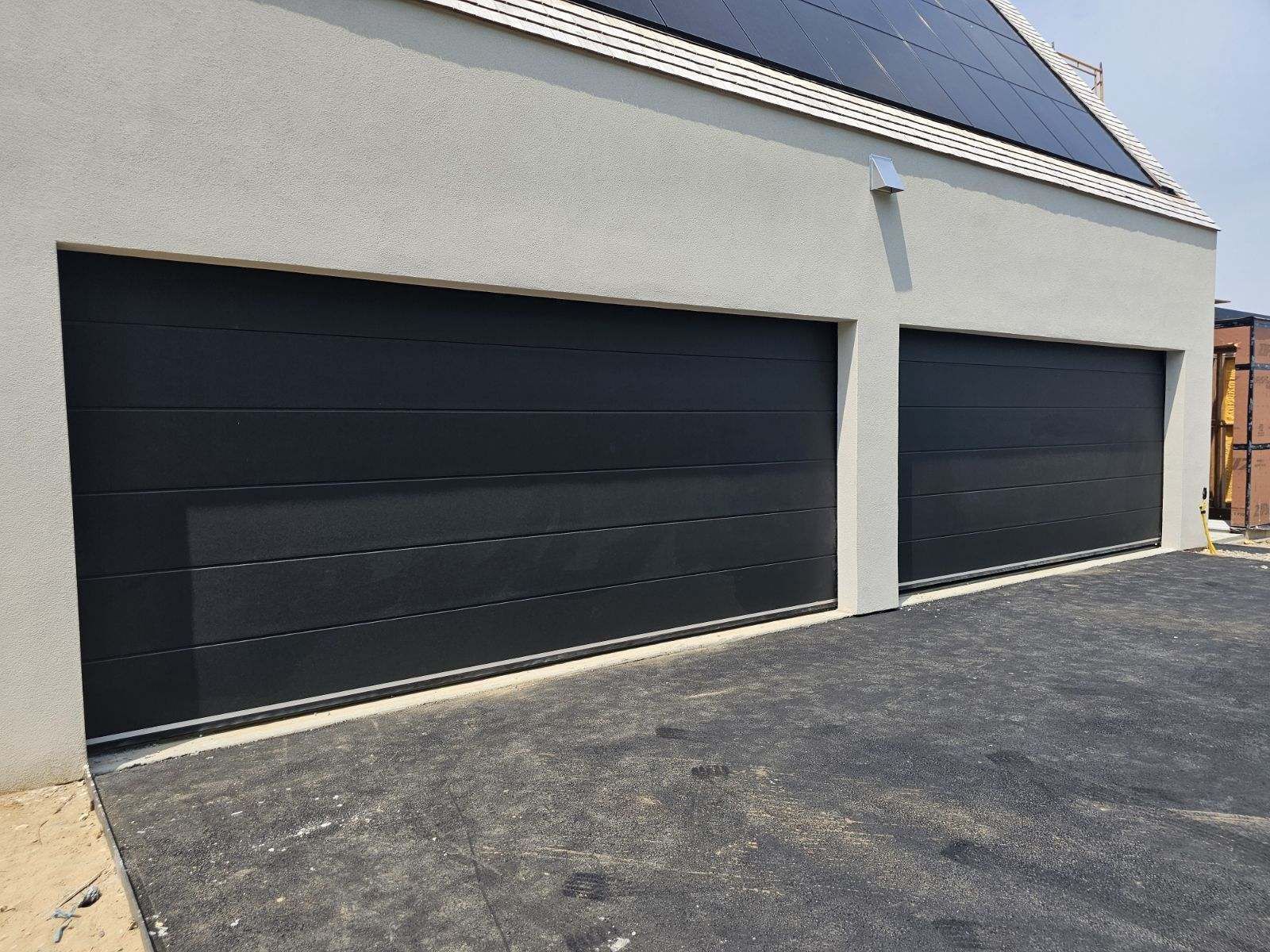 Black garage doors on a light gray building, set on a gravel driveway.