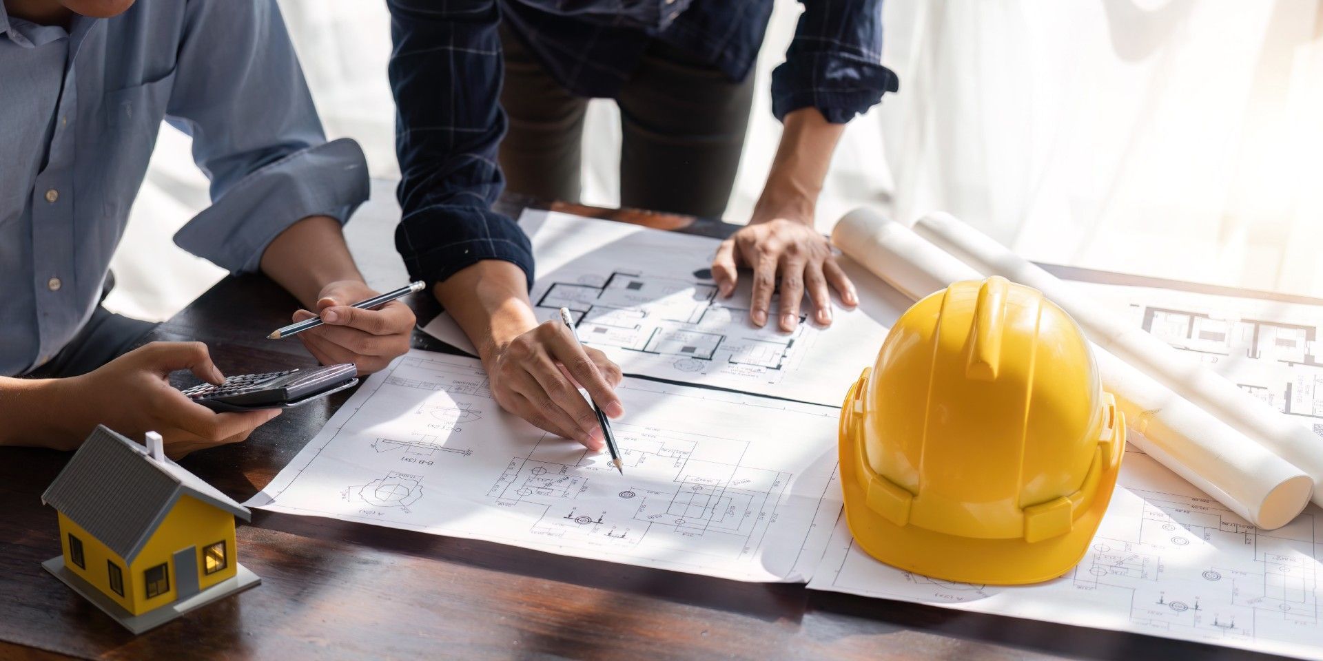 Two people reviewing building plans, with a model house and yellow hard hat on a table.