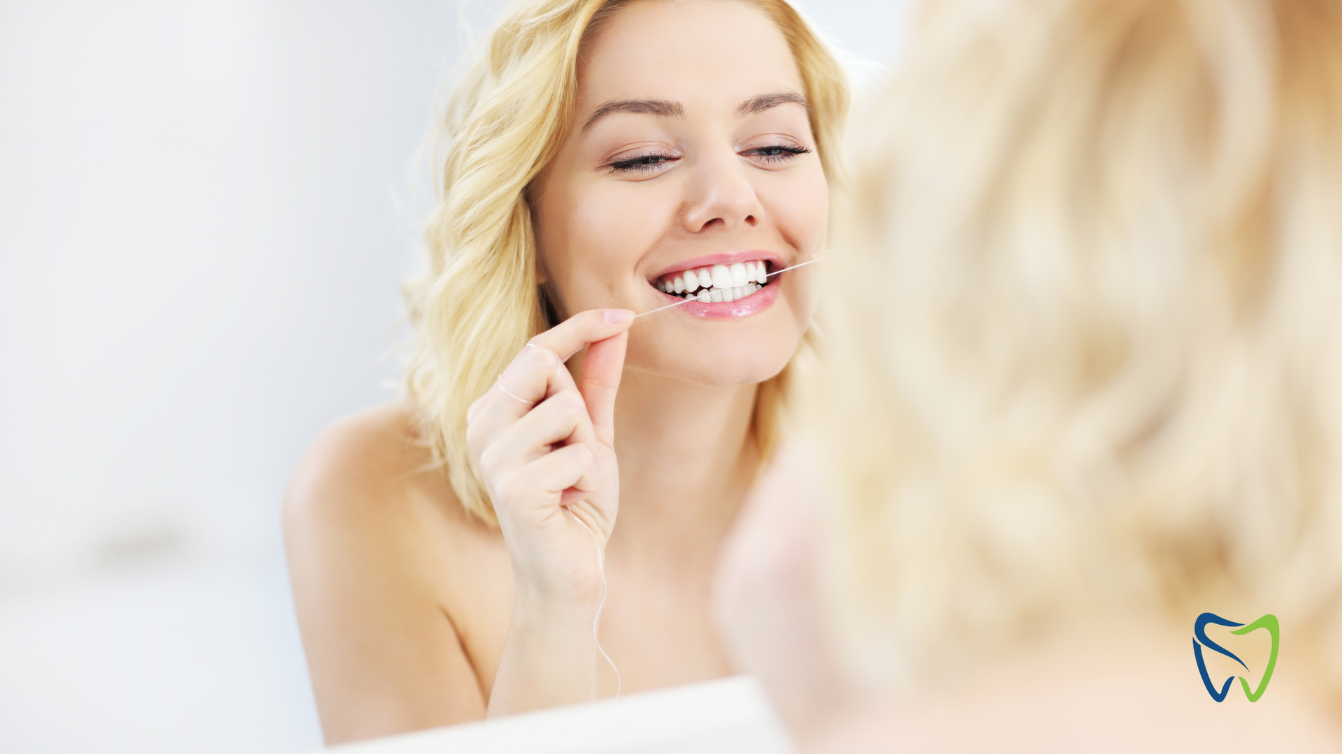 Woman smiling at a mirror, flossing her teeth in a brightly lit bathroom.