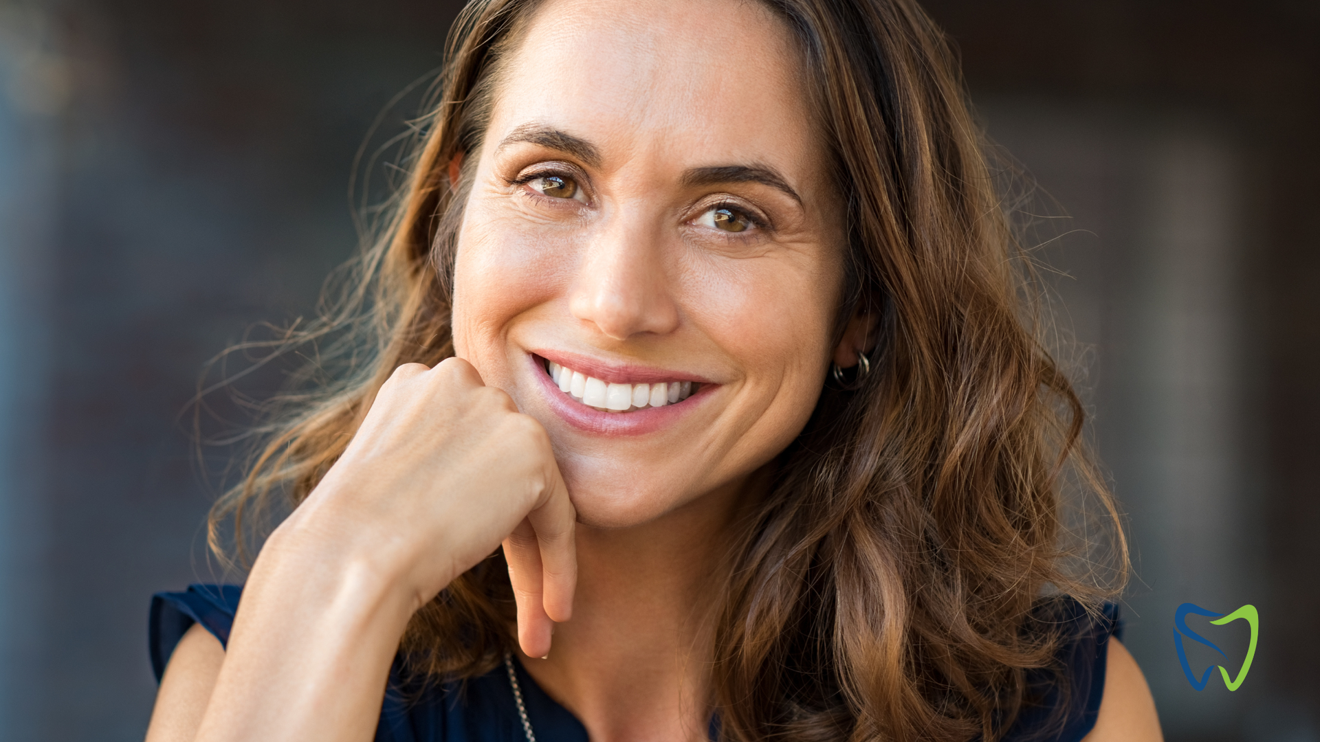 Woman smiling, resting chin on hand. Outdoors, brown hair, dark blue top, bright teeth.