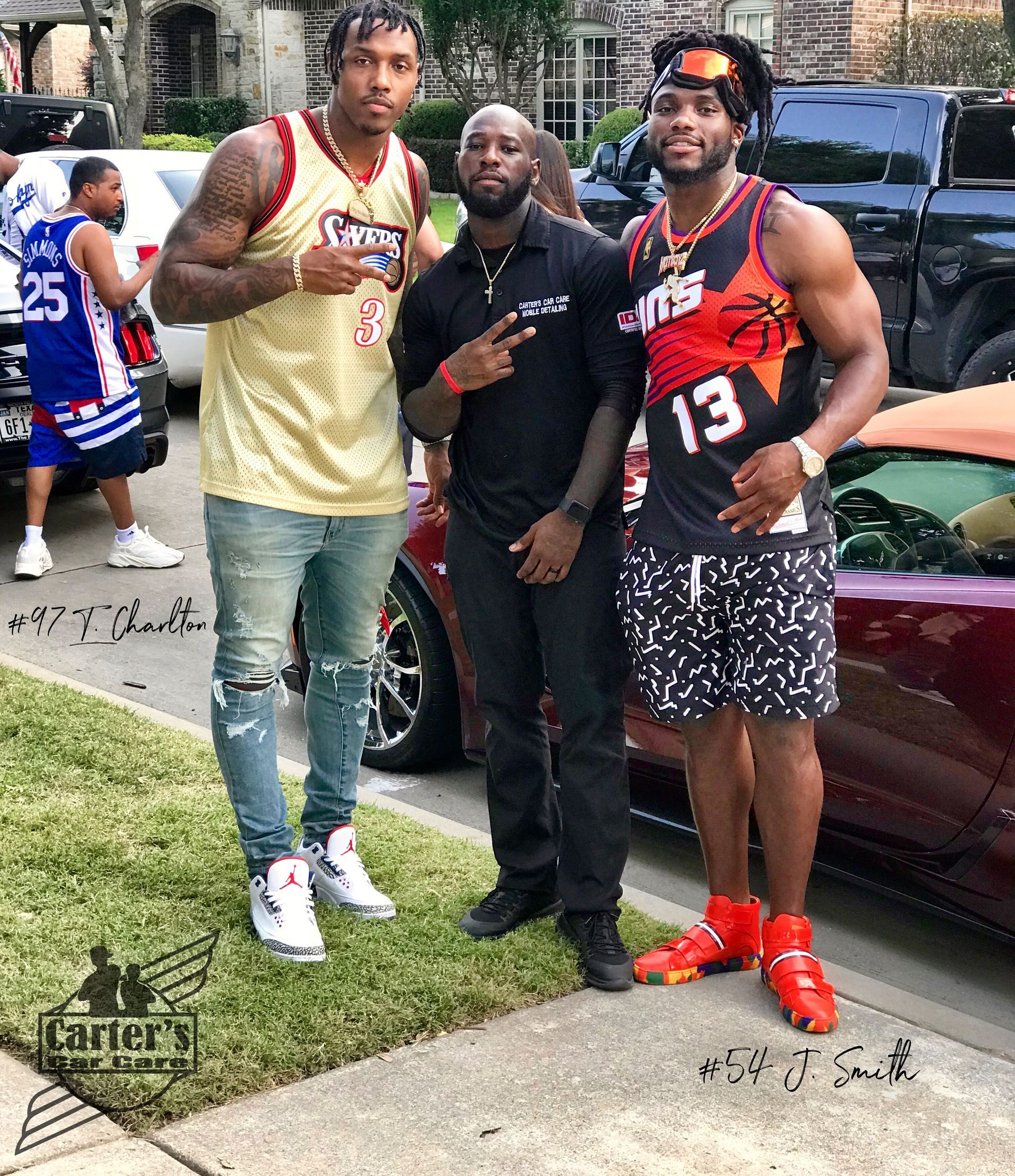 Three men posing outdoors. Two in jerseys, one in black, with cars in the background.