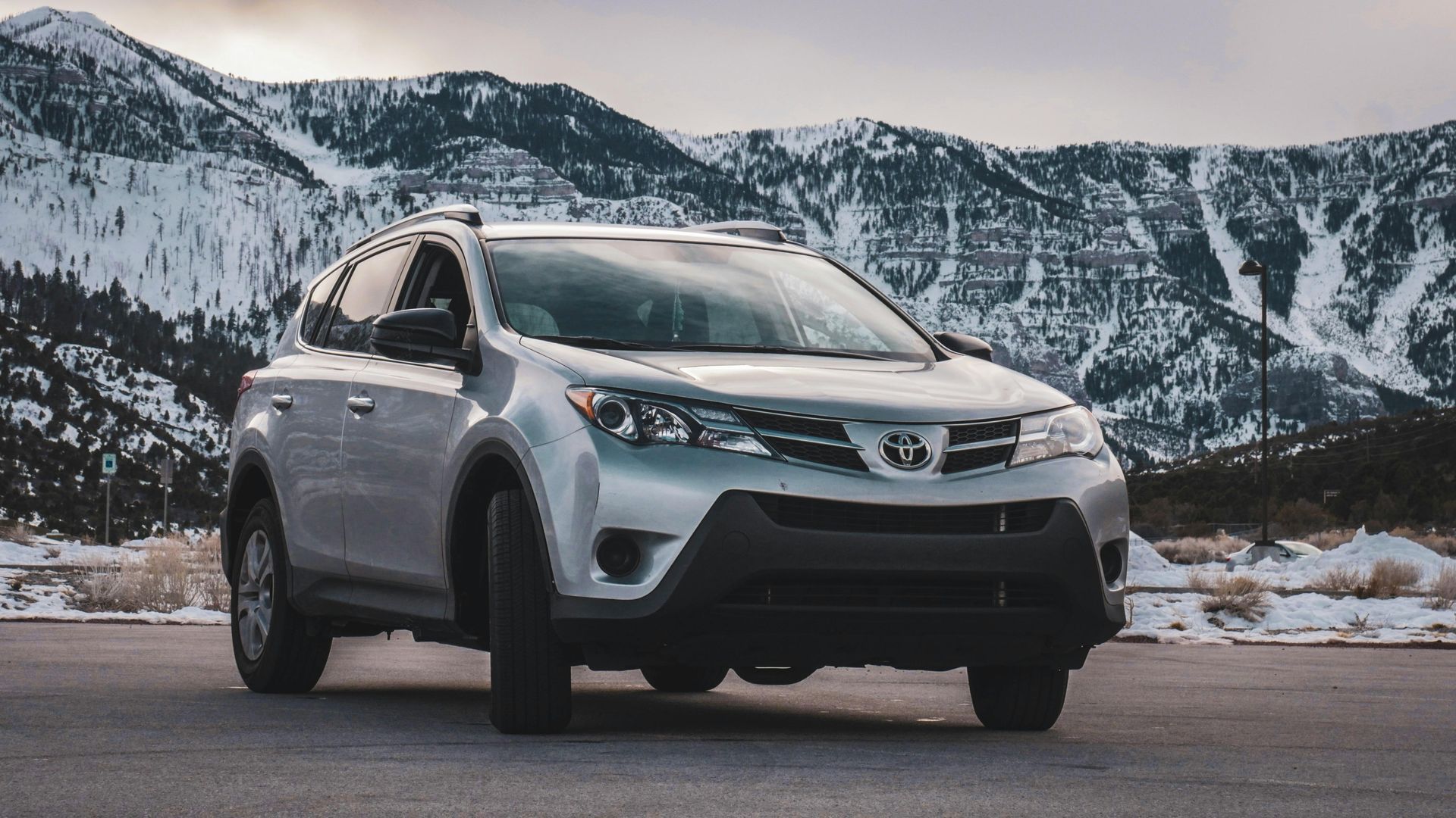 A silver car is parked on the side of the road in front of snow covered mountains.