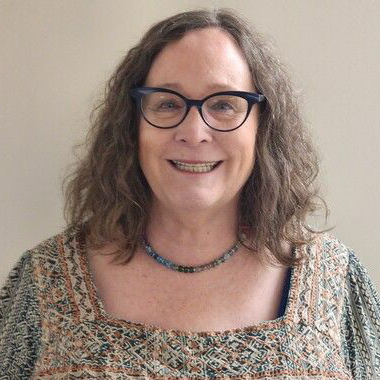 Woman with glasses, smiling, wearing a patterned top and a beaded necklace.