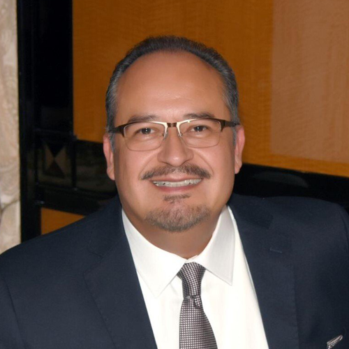 Man in glasses and suit smiles, set against a blurred background.