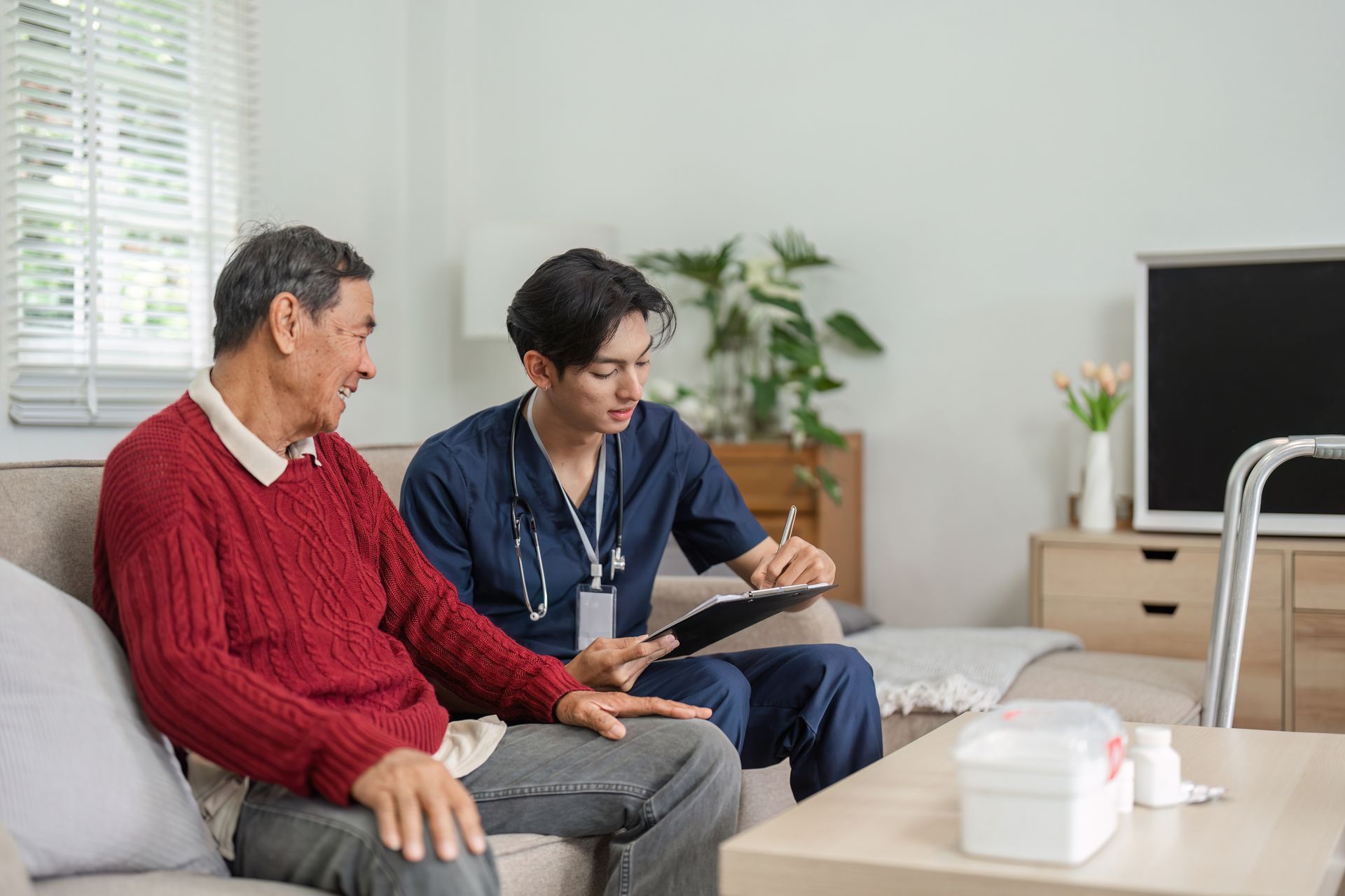 Man in red sweater on couch with caregiver in blue scrubs taking notes. Indoors, daytime.