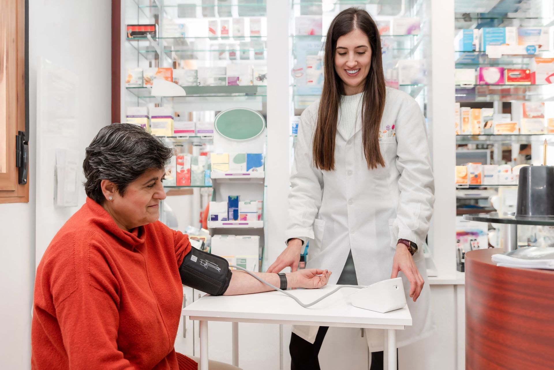 A woman in a red sweater has her blood pressure taken at a pharmacy by a smiling pharmacist.