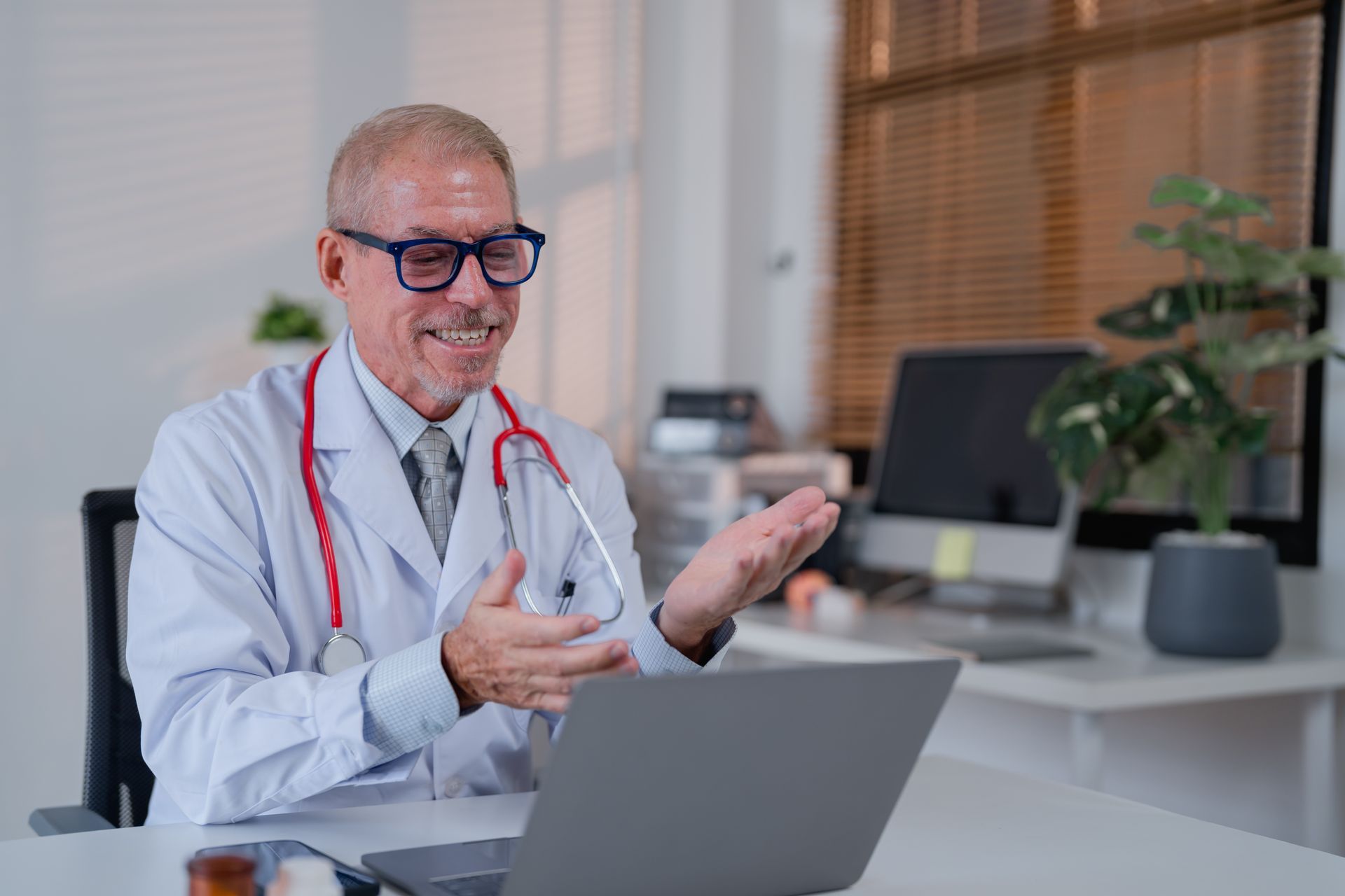 Doctor with glasses smiles while gesturing during a video call on a laptop.