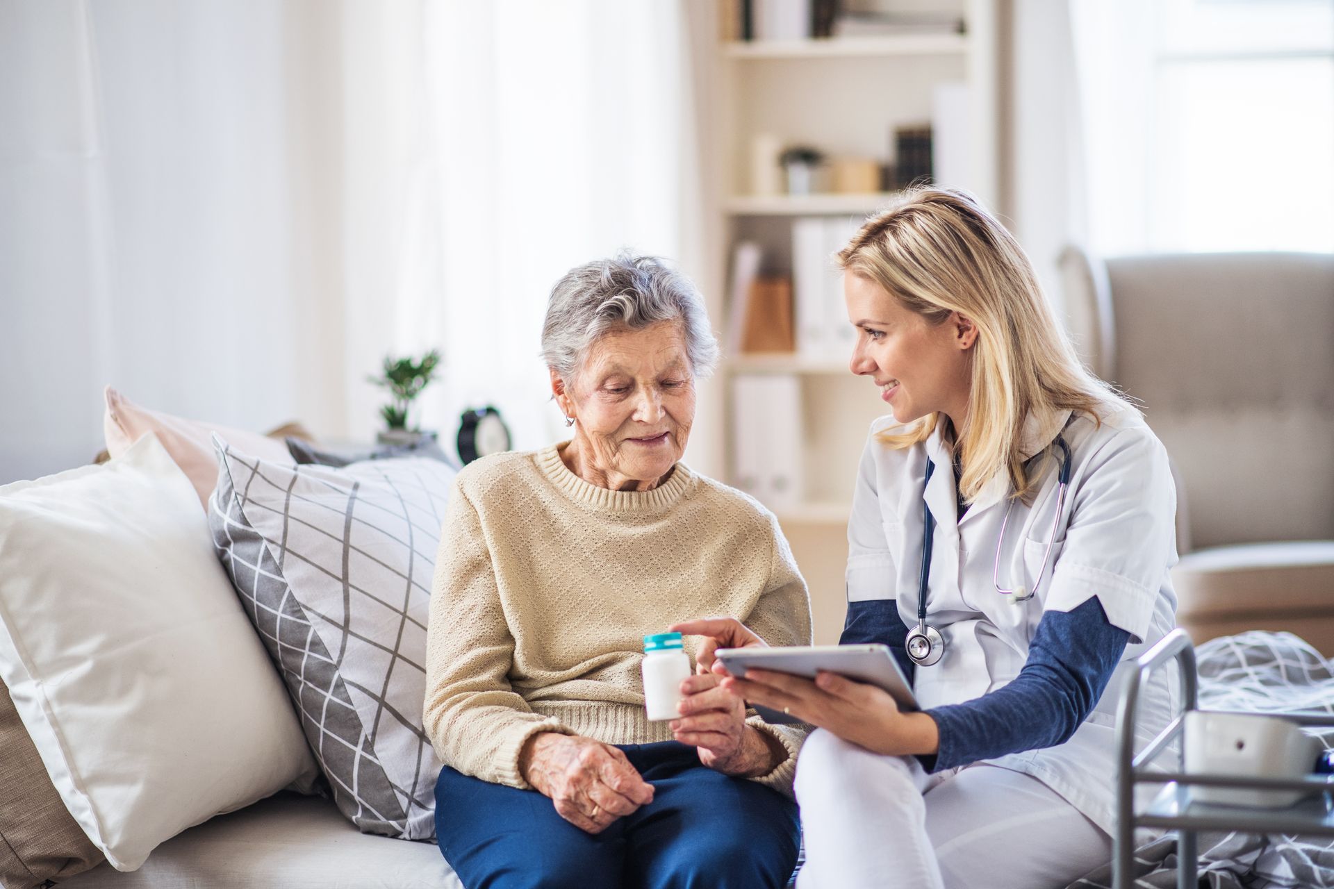 A nurse shows medication to an elderly woman in a home setting. The nurse holds a tablet while smiling.