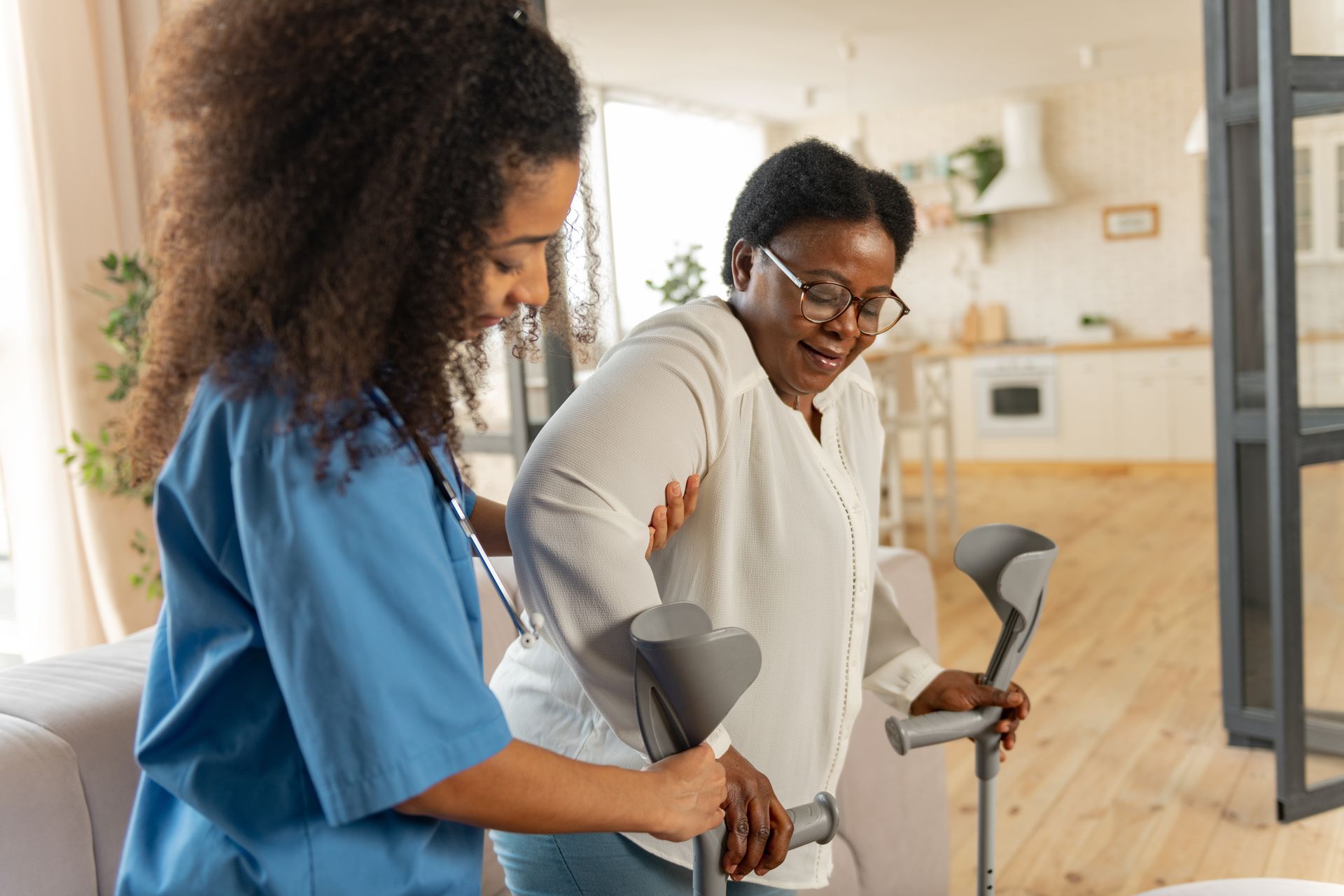 Nurse assisting an older woman using crutches to stand in a home.