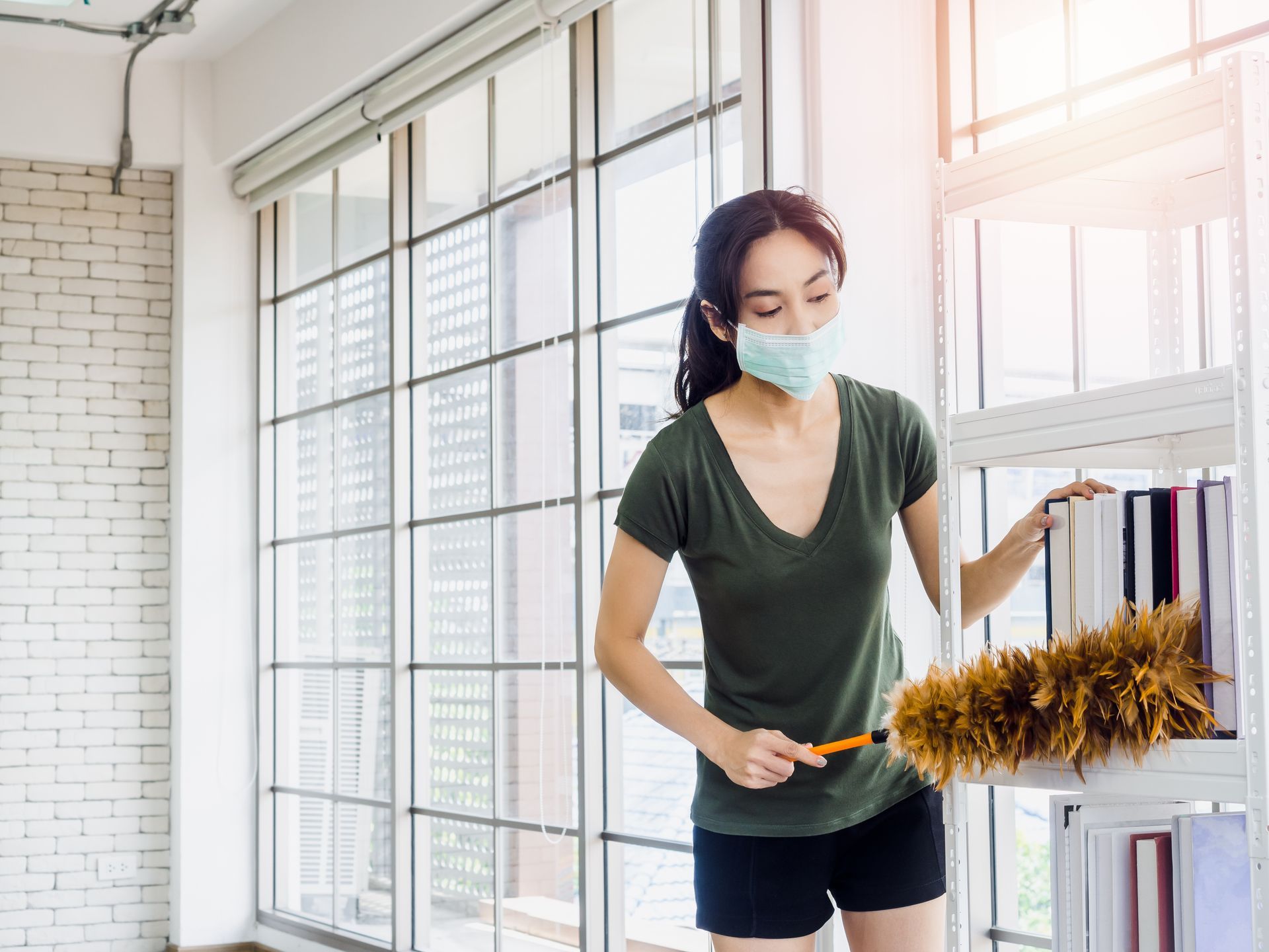 Woman wearing a mask dusting a bookshelf with a feather duster in a bright room with large windows.