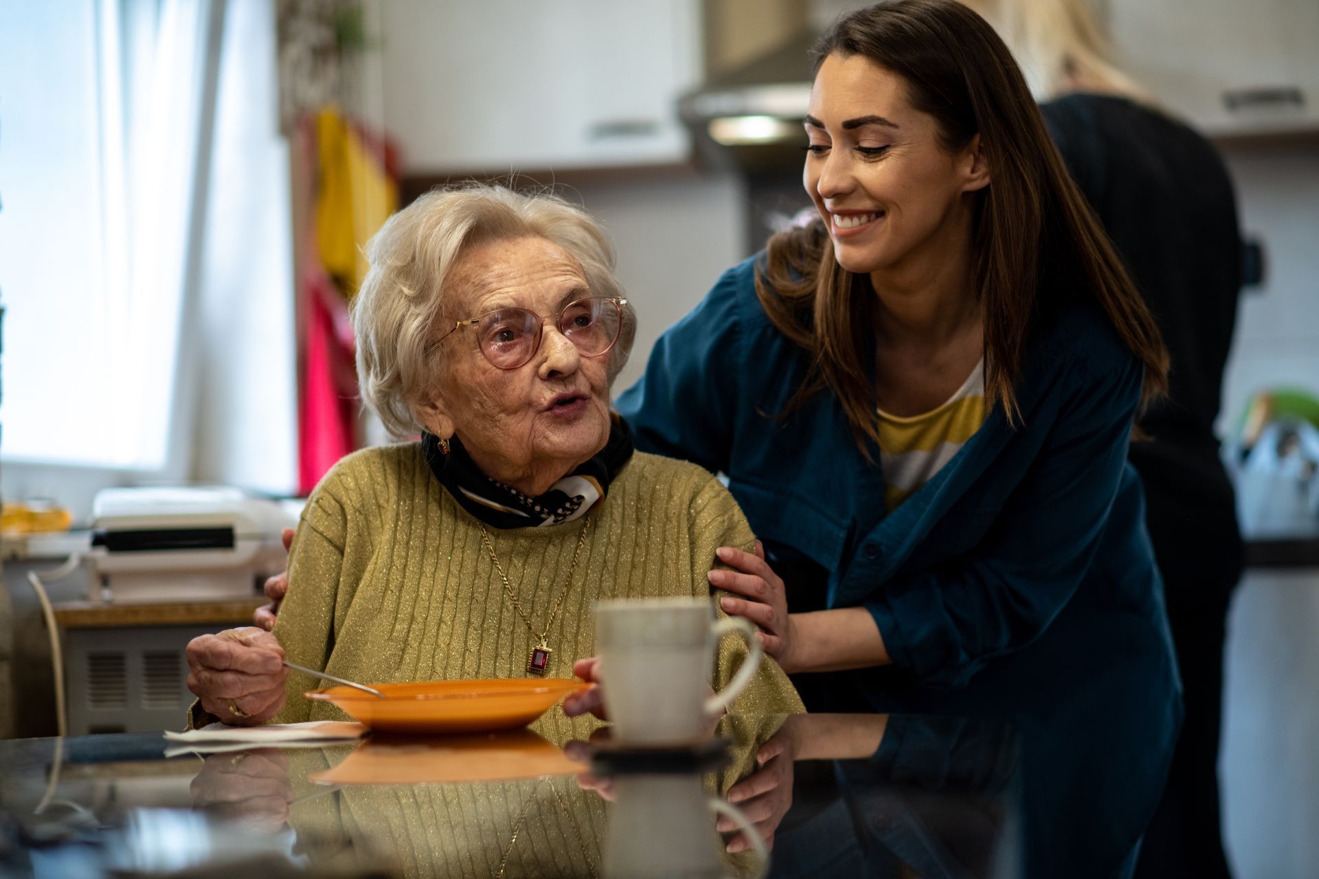 An elderly woman with glasses talks to a young woman in a kitchen. Both are smiling.