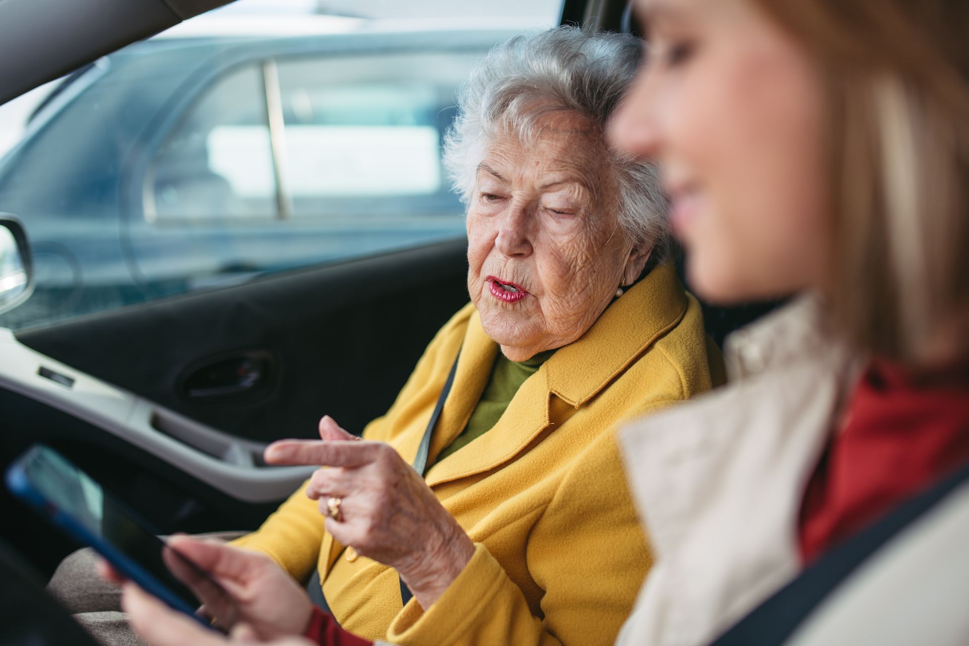 An older woman in a yellow coat points at a phone held by a younger woman in a car.