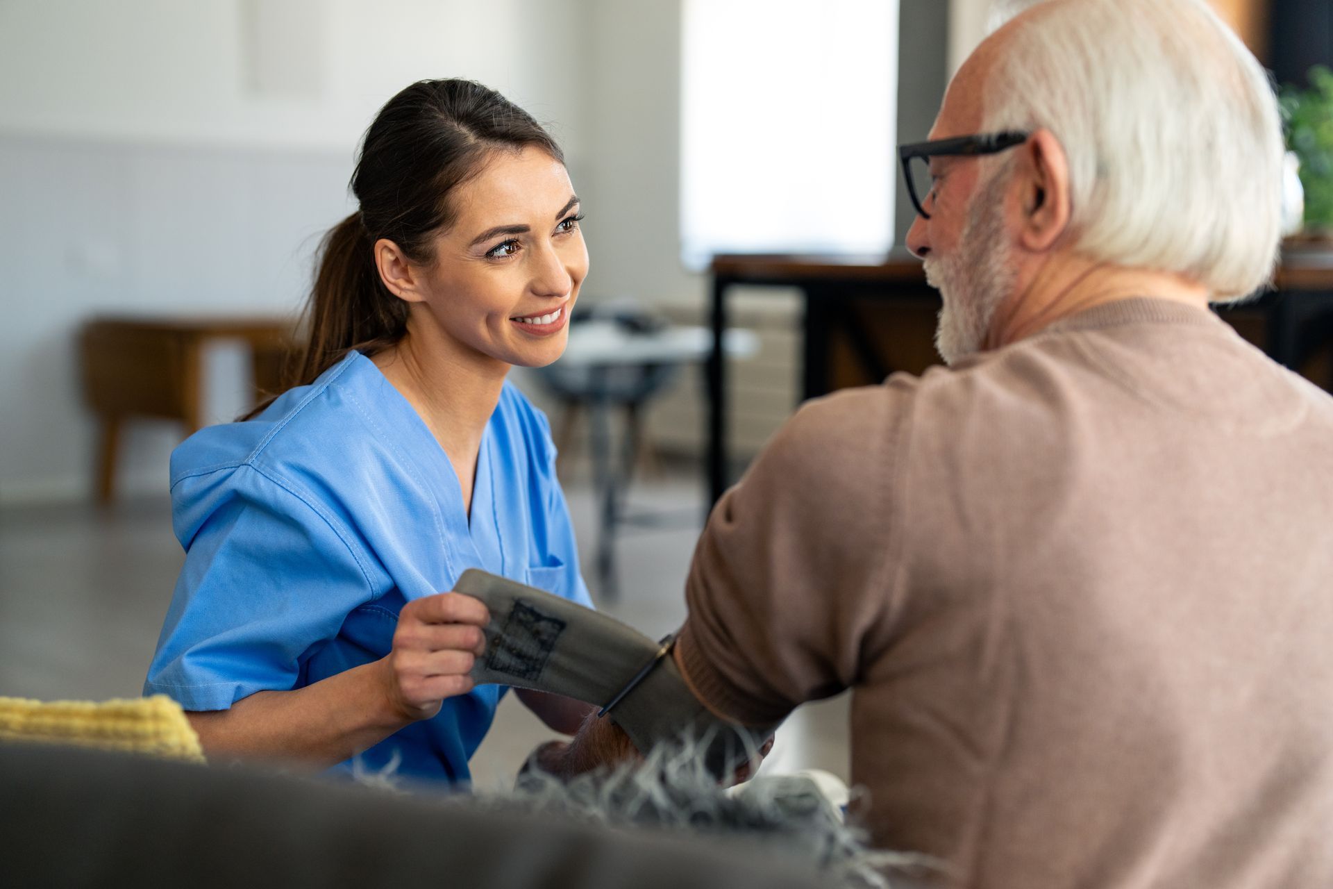 A young woman in blue scrubs checks an elderly man's blood pressure in a home setting.