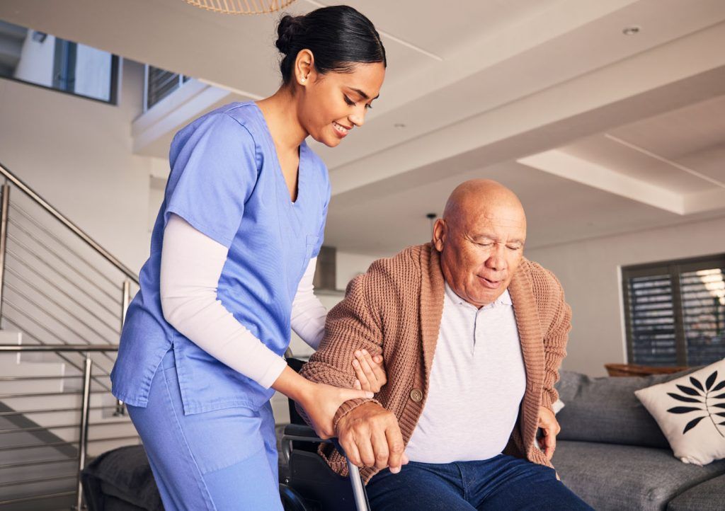 Caregiver in blue scrubs helps a senior man with a cane stand up indoors.