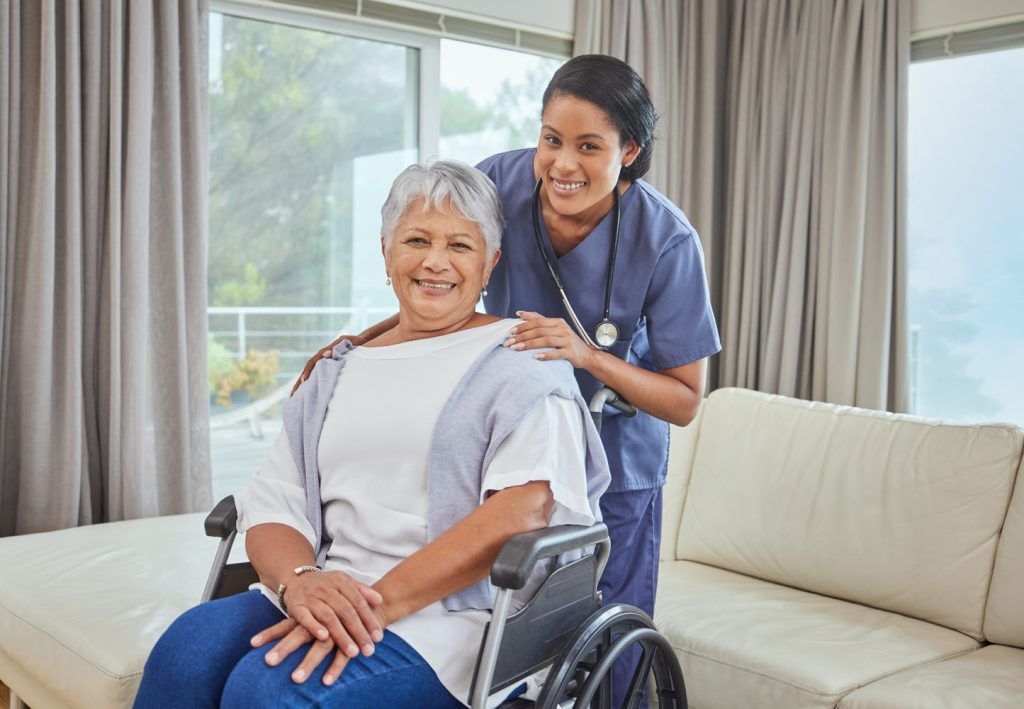 Nurse smiles with senior woman in wheelchair, in a room with a window and a couch.