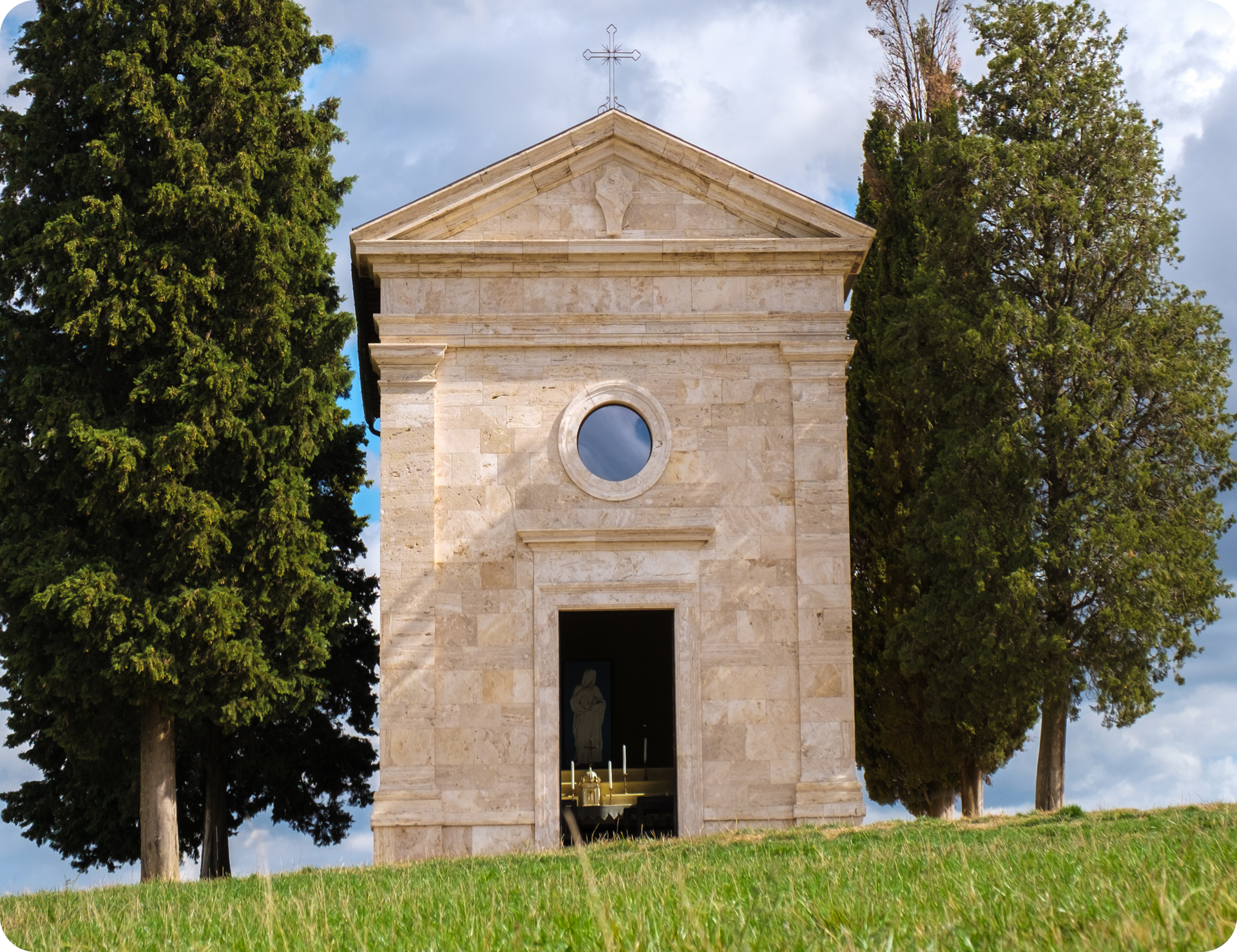 Stone chapel on a grassy hill, framed by tall green trees, under a cloudy sky.