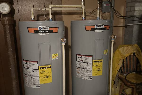 Two grey electric water heaters connected by white pipes in a basement utility area.