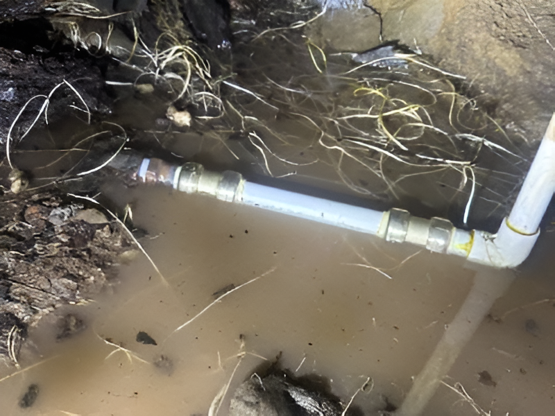 A section of white PVC pipe with metal connectors submerged in muddy water and debris.